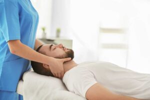 A person gently massaging their jaw and neck while seated at a home desk with a supportive chair.